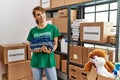 Young caucasian volunteer woman smiling happy holding stack of folded jeans at charity center
