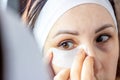 Young caucasian woman applying eye patch mask on her eyelid to avoid wrinkles looking in the mirror. Home skin care procedure