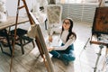 Young caucasian woman artist sitting on a floor holding paintbrush and draws on canvas working on a home studio Royalty Free Stock Photo