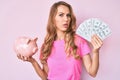 Young caucasian woman with blond hair holding dollars and piggy bank clueless and confused expression Royalty Free Stock Photo