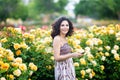 Young Caucasian woman with dark curly hair near yellow rose bush in a rose garden looking to the camera and smiling holding black Royalty Free Stock Photo