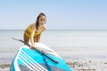 Young Caucasian woman dragging a paddle surfboard from the beach to the sea with gesture