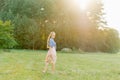 Young Caucasian woman or girl in a summer dress and a denim shirt is walking on the grass, holding shoes in her hands, relaxing Royalty Free Stock Photo