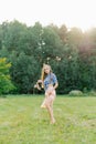 Young Caucasian woman or girl in a summer dress and a denim shirt is walking on the grass, holding shoes in her hands, relaxing Royalty Free Stock Photo