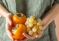 Young Caucasian woman in gray linen shirt holds in hand bunch of ripe juicy white grapes persimmons. Autumn harvest healthy life