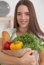 Young caucasian woman in a green apron is holding paper bag full of vegetables and fruits while smiling in kitchen Royalty Free Stock Photo