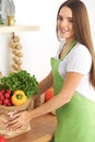 Young caucasian woman in a green apron is holding paper bag full of vegetables and fruits while smiling in kitchen Royalty Free Stock Photo