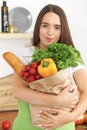 Young caucasian woman in a green apron is holding paper bag full of vegetables and fruits while smiling in kitchen Royalty Free Stock Photo