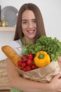 Young caucasian woman in a green apron is holding paper bag full of vegetables and fruits while smiling in kitchen Royalty Free Stock Photo