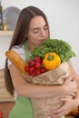 Young caucasian woman in a green apron is holding paper bag full of vegetables and fruits while smiling in kitchen Royalty Free Stock Photo