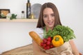 Young caucasian woman in a green apron is holding paper bag full of vegetables and fruits while smiling in kitchen Royalty Free Stock Photo