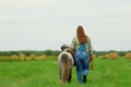 The young Caucasian woman and her gray pony are walking on the grass in rural, the back view