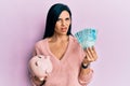 Young caucasian woman holding brazil real banknotes and piggy bank in shock face, looking skeptical and sarcastic, surprised with