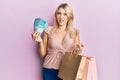 Young caucasian woman holding brazil real banknotes and shopping bags in shock face, looking skeptical and sarcastic, surprised