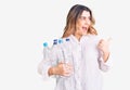 Young caucasian woman holding recycling plastic bottles pointing thumb up to the side smiling happy with open mouth Royalty Free Stock Photo