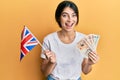 Young caucasian woman holding uk flag and pounds banknotes smiling and laughing hard out loud because funny crazy joke