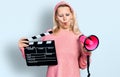 Young caucasian woman holding video film clapboard and megaphone making fish face with mouth and squinting eyes, crazy and comical Royalty Free Stock Photo