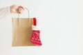 Young caucasian woman holds in hand blank craft paper bag with hanging knitted red Christmas gift stocking on white wall Royalty Free Stock Photo