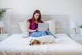 Young caucasian woman at home working on laptop with cute jack russell dog resting on bed. Home office, Pets, love and relax. Royalty Free Stock Photo