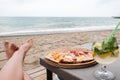 Young Caucasian woman lying at the beach eating pizza and drinking cocktail. Legs, sea in background. Careless holidays at all