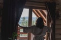 Young caucasian woman opening curtains in a wooden chalet cabin in the mountains. Comfortable rest in a log house in nature