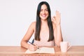 Young caucasian woman sitting at the desk writing book drinking coffee doing ok sign with fingers, smiling friendly gesturing Royalty Free Stock Photo