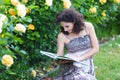 Young Caucasian woman sitting on green grass near yellow roses bush in a rose garden, reading a book, looking to the book Royalty Free Stock Photo