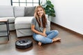 Young caucasian woman sitting at home by vacuum robot looking positive and happy standing and smiling with a confident smile Royalty Free Stock Photo