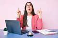 Young caucasian woman sitting at the recepcionist desk working using laptop amazed and surprised looking up and pointing with