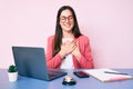 Young caucasian woman sitting at the recepcionist desk working using laptop smiling with hands on chest with closed eyes and