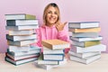 Young caucasian woman sitting on the table with books pointing to the back behind with hand and thumbs up, smiling confident Royalty Free Stock Photo