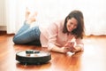 Young caucasian woman smiling in the living room and using automatic vacuum cleaner to clean the floor, controlling smart machine