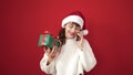 Young caucasian woman smiling wearing christmas hat holding gift speaking on the phone over isolated red background Royalty Free Stock Photo