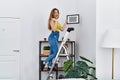 Young caucasian woman on stairs hanging picture on the wall screaming proud, celebrating victory and success very excited with Royalty Free Stock Photo