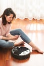 Young caucasian woman using automatic vacuum cleaner to clean the floor, controlling smart machine housework robot