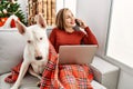 Young caucasian woman using laptop and talking on the smartphone sitting with dog by christmas tree at home Royalty Free Stock Photo