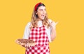 Young caucasian woman wearing apron holding pizza pointing thumb up to the side smiling happy with open mouth Royalty Free Stock Photo