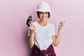 Young caucasian woman wearing architect hardhat using drill smiling happy pointing with hand and finger to the side Royalty Free Stock Photo