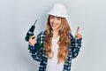 Young caucasian woman wearing architect hardhat using drill smiling with an idea or question pointing finger with happy face, Royalty Free Stock Photo