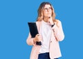 Young caucasian woman wearing business clothes and glasses holding binder serious face thinking about question with hand on chin, Royalty Free Stock Photo