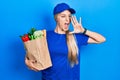 Young caucasian woman wearing courier uniform with groceries from supermarket shouting and screaming loud to side with hand on Royalty Free Stock Photo