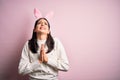Young caucasian woman wearing cute easter rabbit ears over pink isolated background begging and praying with hands together with