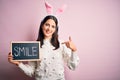 Young caucasian woman wearing easter rabbit ears and holding blackboard with smile word with surprise face pointing finger to