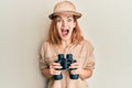 Young caucasian woman wearing explorer hat looking through binoculars celebrating crazy and amazed for success with open eyes