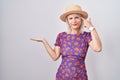Young caucasian woman wearing flowers dress and summer hat confused and annoyed with open palm showing copy space and pointing