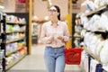 Young Caucasian woman wearing glasses holds grocery basket, holds list and selects bed linen and pillow. Shopping and