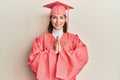 Young caucasian woman wearing graduation cap and ceremony robe praying with hands together asking for forgiveness smiling