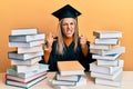 Young caucasian woman wearing graduation ceremony robe sitting on the table angry and mad raising fists frustrated and furious Royalty Free Stock Photo