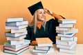 Young caucasian woman wearing graduation ceremony robe sitting on the table smiling confident touching hair with hand up gesture, Royalty Free Stock Photo