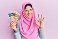 Young caucasian woman wearing islamic hijab holding canadian dollars banknotes doing ok sign with fingers, smiling friendly